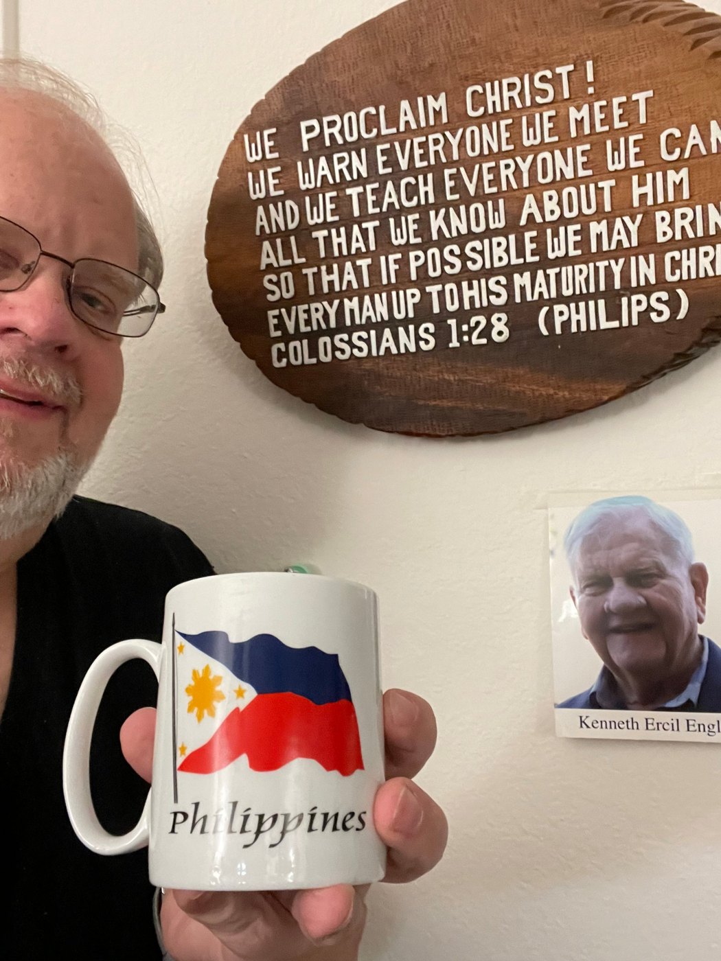 Kevin Engle holding a Philippines mug beside his father’s Colossians 1:28 plaque and memorial photograph